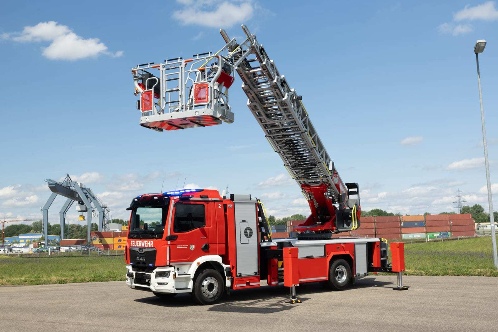 A red Rosenbauer L32A-XS electric aerial ladder on a MAN chassis with its ladder and rescue basket raised against a blue sky.