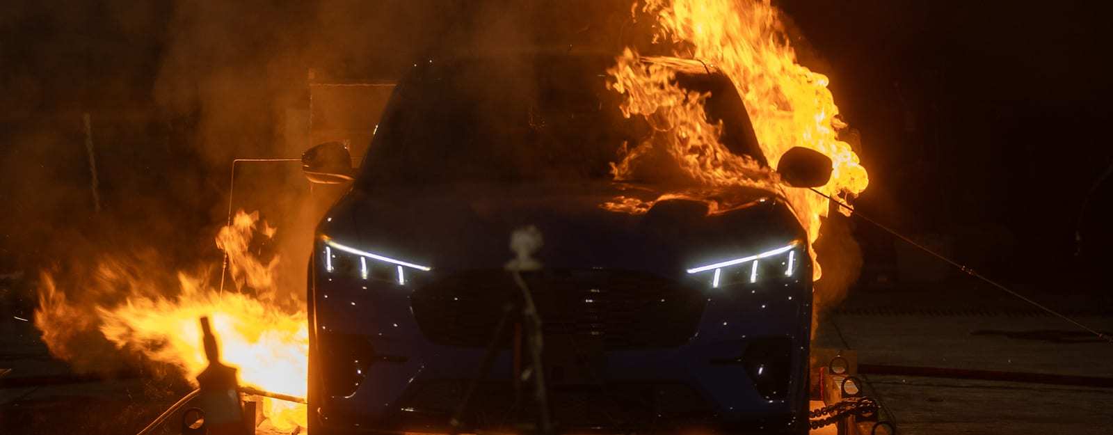 A dark blue electric sport utility vehicle (SUV) with its headlights on faces the camera in a darkened test lab, with flames rising over one side of the windshield and a burner flame beside the front bumper on the other side.