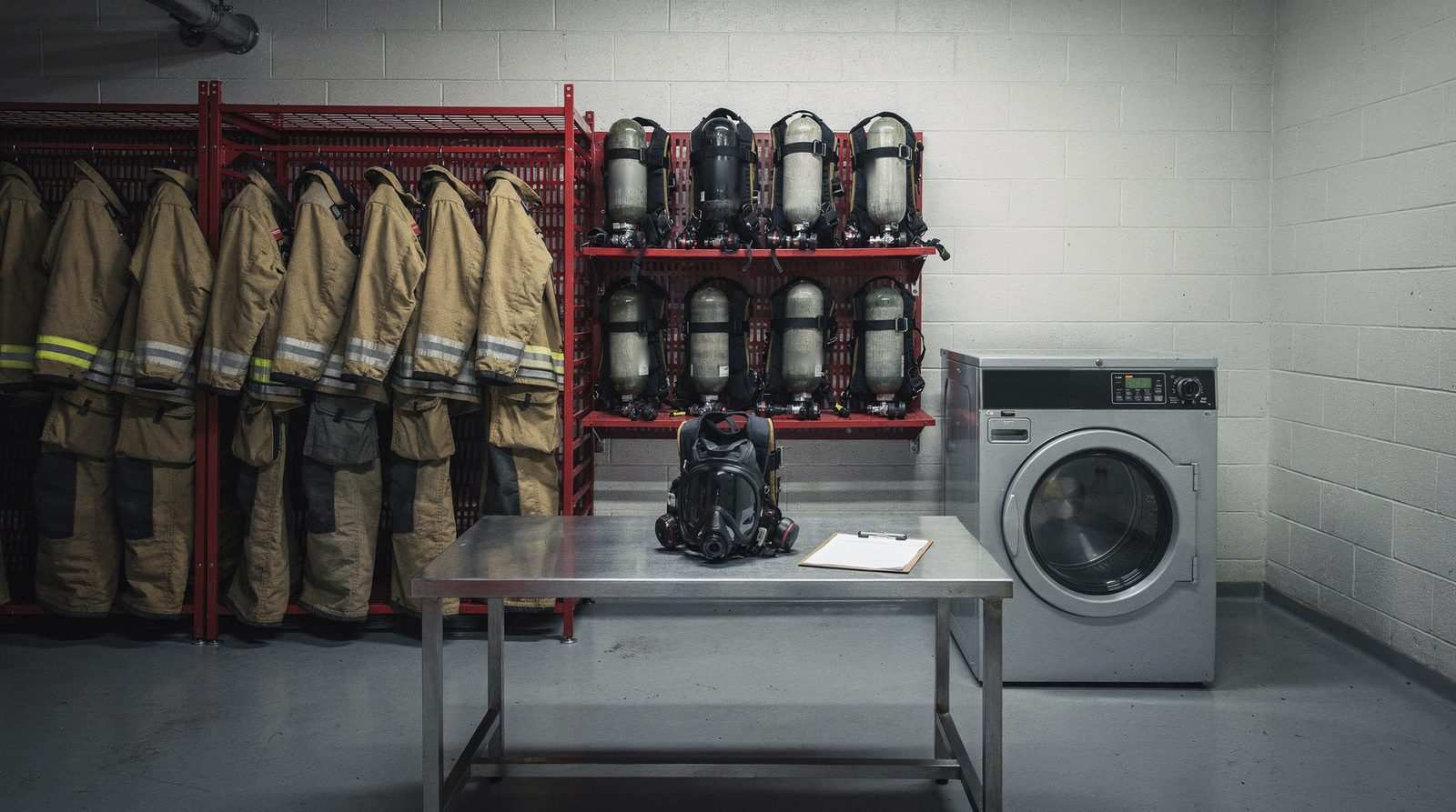 A fire-station gear room with turnout coats on open racks, breathing apparatus on wall shelves, a facepiece and a blank clipboard on a steel bench, and a front-loading washer.