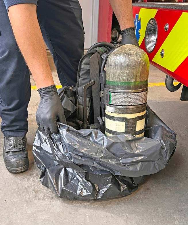 A firefighter in gloves lowers an SCBA with its cylinder into a large black plastic bag.