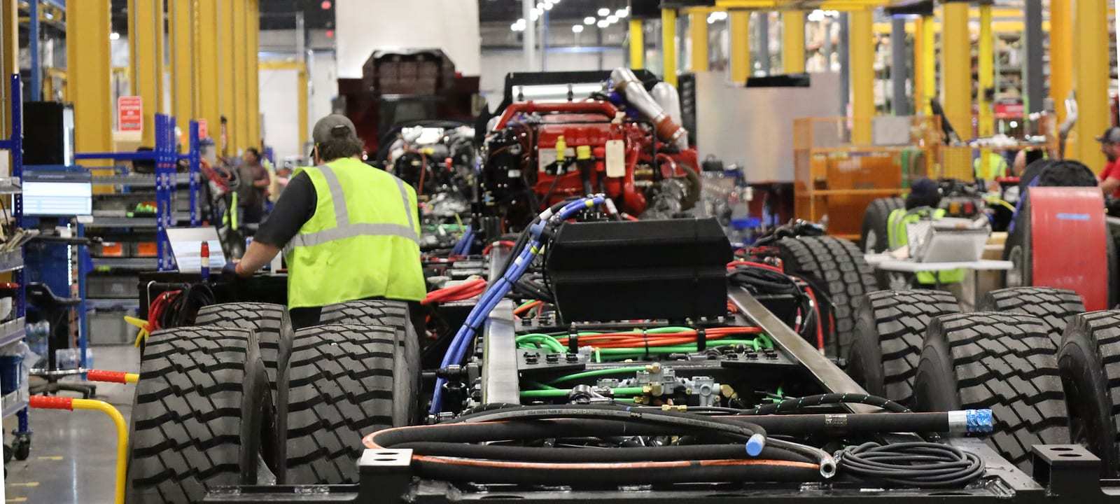 Fire truck chassis with engines, wiring harnesses and tires mounted move down an assembly line in a Pierce plant while a worker in a high-visibility vest works at a station.