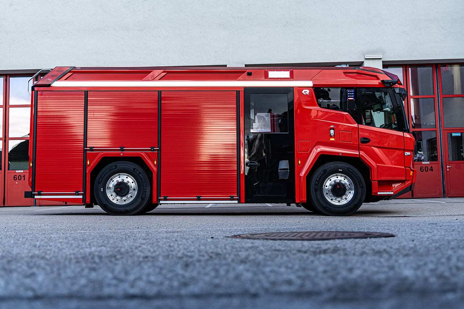 Side view of a red Rosenbauer RT electric fire engine without department markings, parked in front of red fire station bay doors numbered 601 and 604.